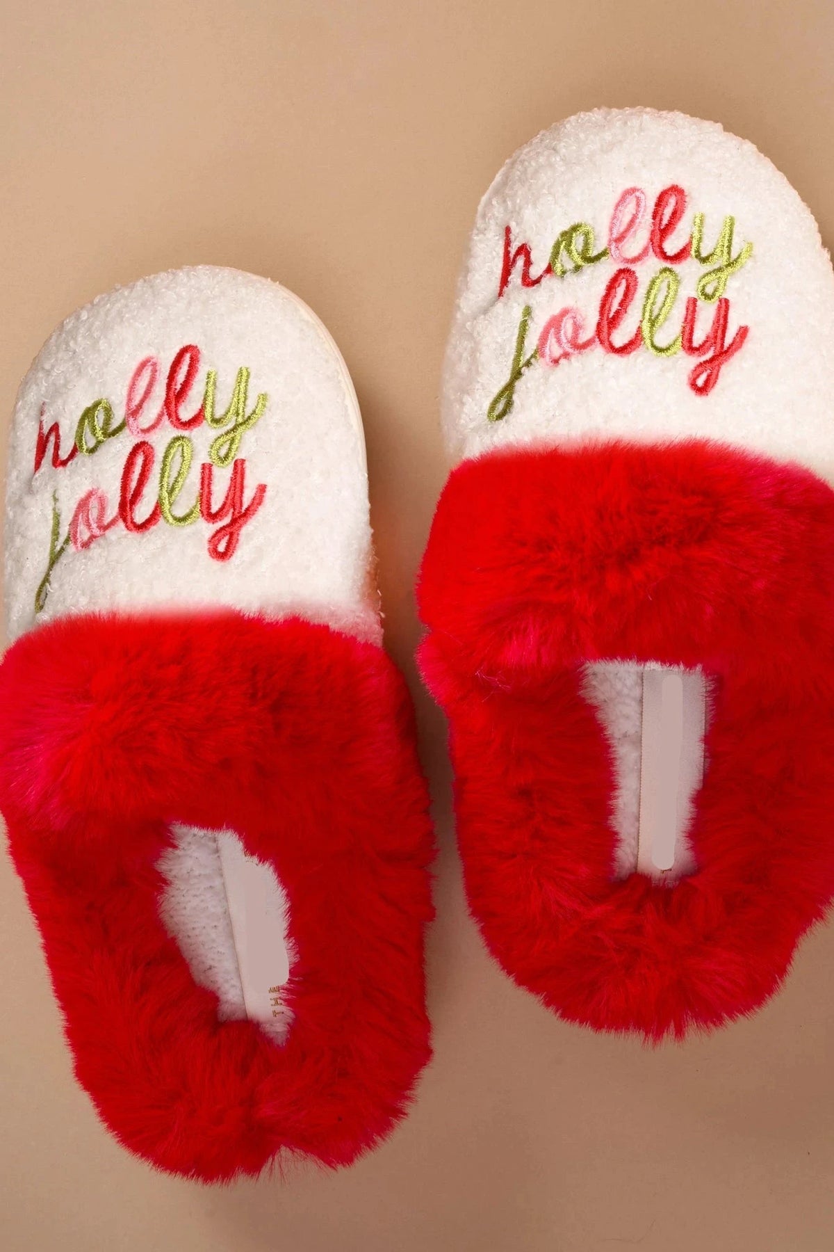 A pair of white and red fuzzy slippers with 'holly jolly' embroidered in green and red on the top, shown on a solid beige background.