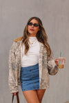 Woman in a camouflage jacket and denim skirt holding a pumpkin spice latte against a gray background