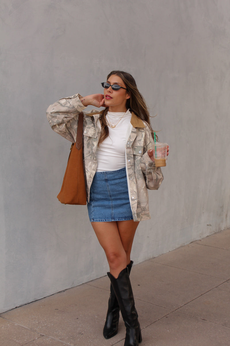 Woman in a camouflage jacket, white top, blue skirt, and black boots standing against a gray wall.