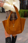Brown suede handbag held by a person wearing a white sweater and plaid skirt.