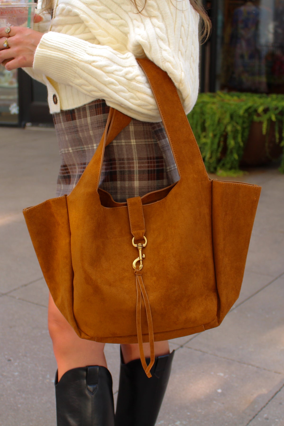 Brown suede handbag held by a person wearing a white sweater and plaid skirt.