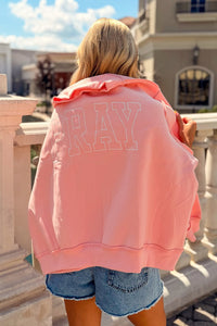 A woman viewed from behind wearing a pink sweatshirt with ‘PRAY' outlined in white across the back, paired with denim shorts, standing outdoors on a balcony.