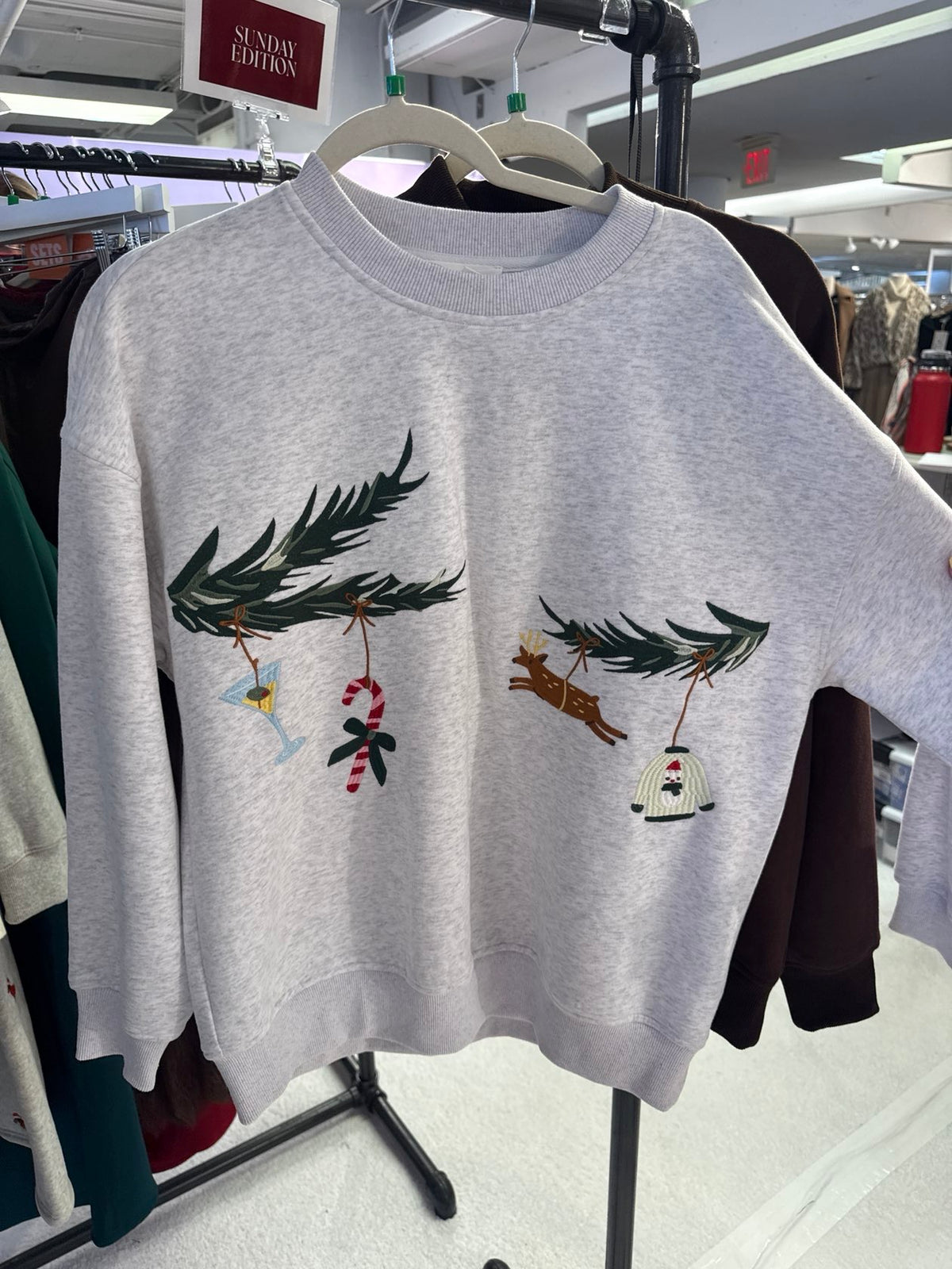 A light gray crewneck sweatshirt hanging on a hanger in a retail store. The sweatshirt features embroidered Christmas tree branches with hanging ornaments, including a martini glass, candy cane, reindeer, and a small white house.