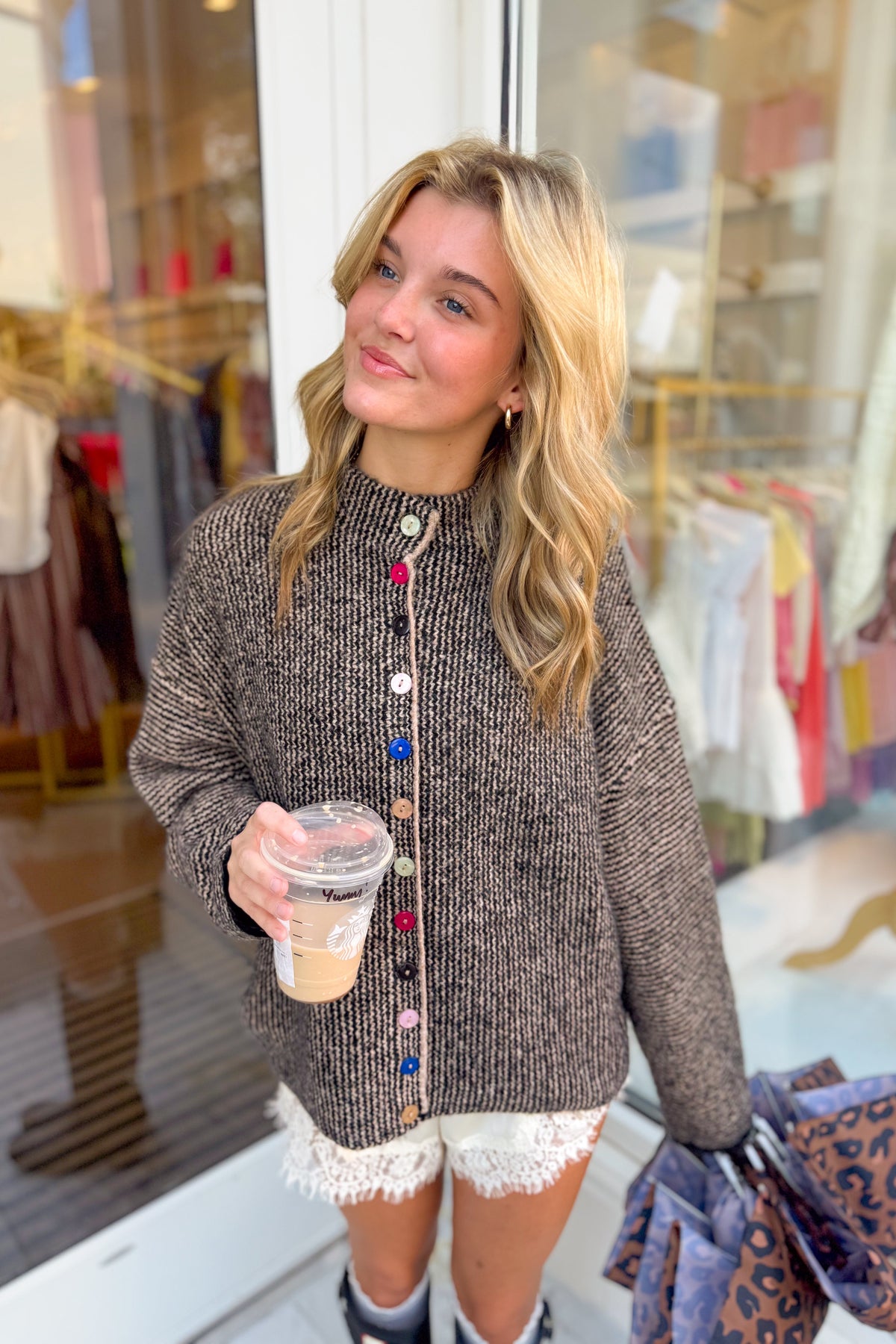 A blonde woman stands in front of a clothing store window wearing a brown ribbed knit button-up sweater with colorful buttons, white lace-trim shorts, and holding an iced coffee.