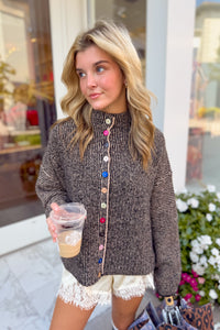 A woman wearing a brown textured knit mock-neck sweater with colorful decorative buttons down the front, holding a coffee cup in front of a storefront.