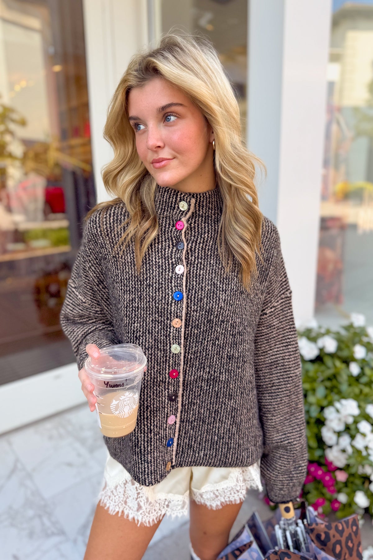 A woman wearing a brown textured knit mock-neck sweater with colorful decorative buttons down the front, holding a coffee cup in front of a storefront.