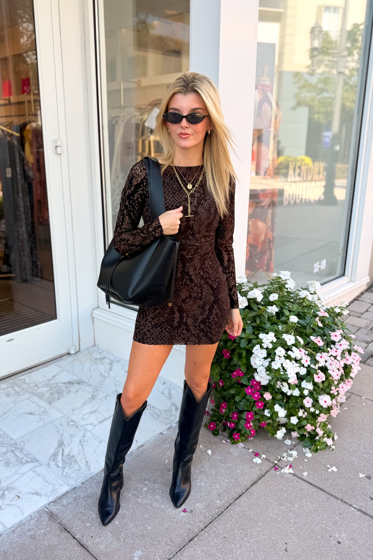 A woman stands on a sidewalk in front of a storefront, wearing a dark brown lace long-sleeve mini dress, black pointed-toe knee-high boots, and carrying a large black shoulder bag.