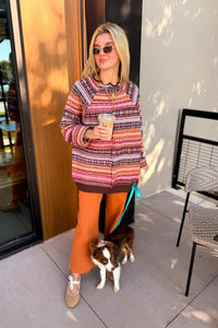 A woman stands on a patio wearing a multicolor patterned knit sweater, rust-orange pants, and tan loafers, holding a drink while standing next to a small dog.