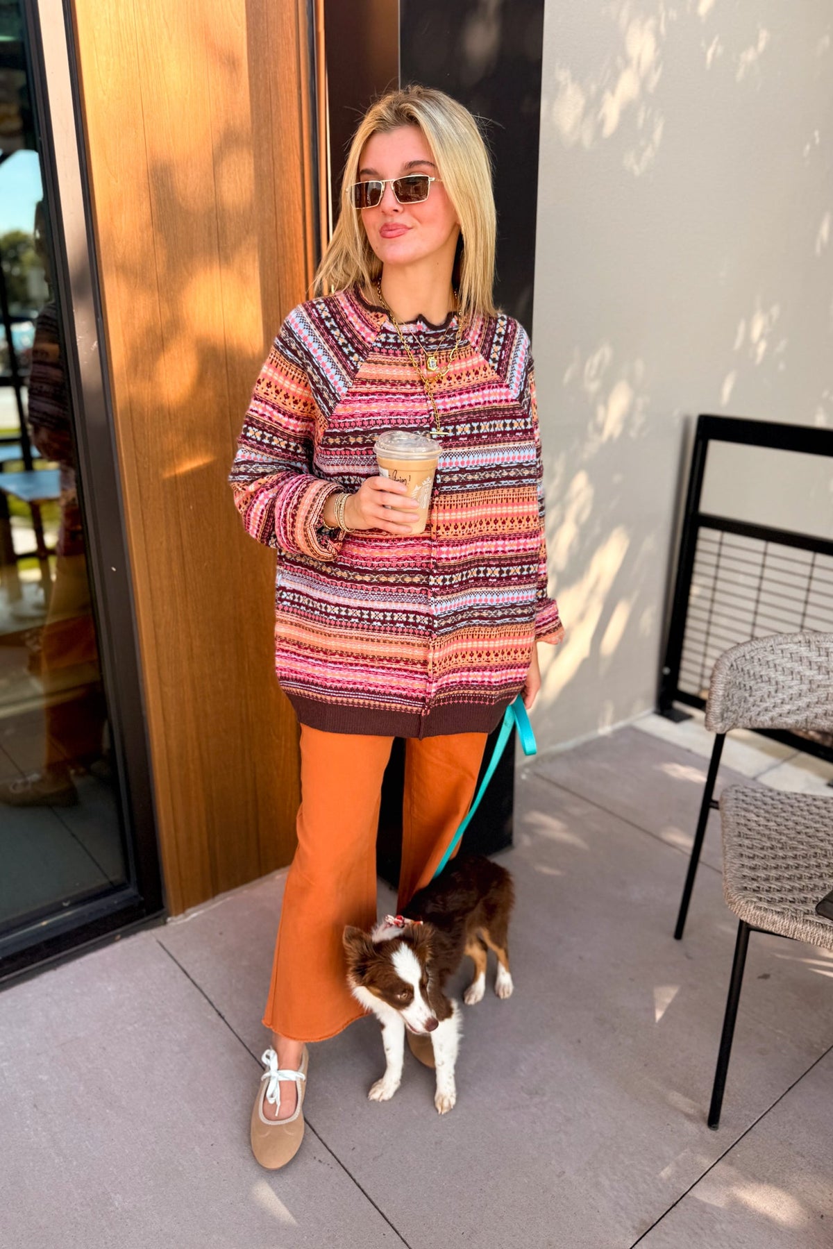A woman stands on a patio wearing a multicolor patterned knit sweater, rust-orange pants, and tan loafers, holding a drink while standing next to a small dog.