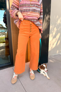 A person wearing bright orange wide-leg cropped pants with a button fly, styled with a multicolor patterned knit sweater and beige flat shoes, standing outdoors next to a brown and white dog on a teal leash.