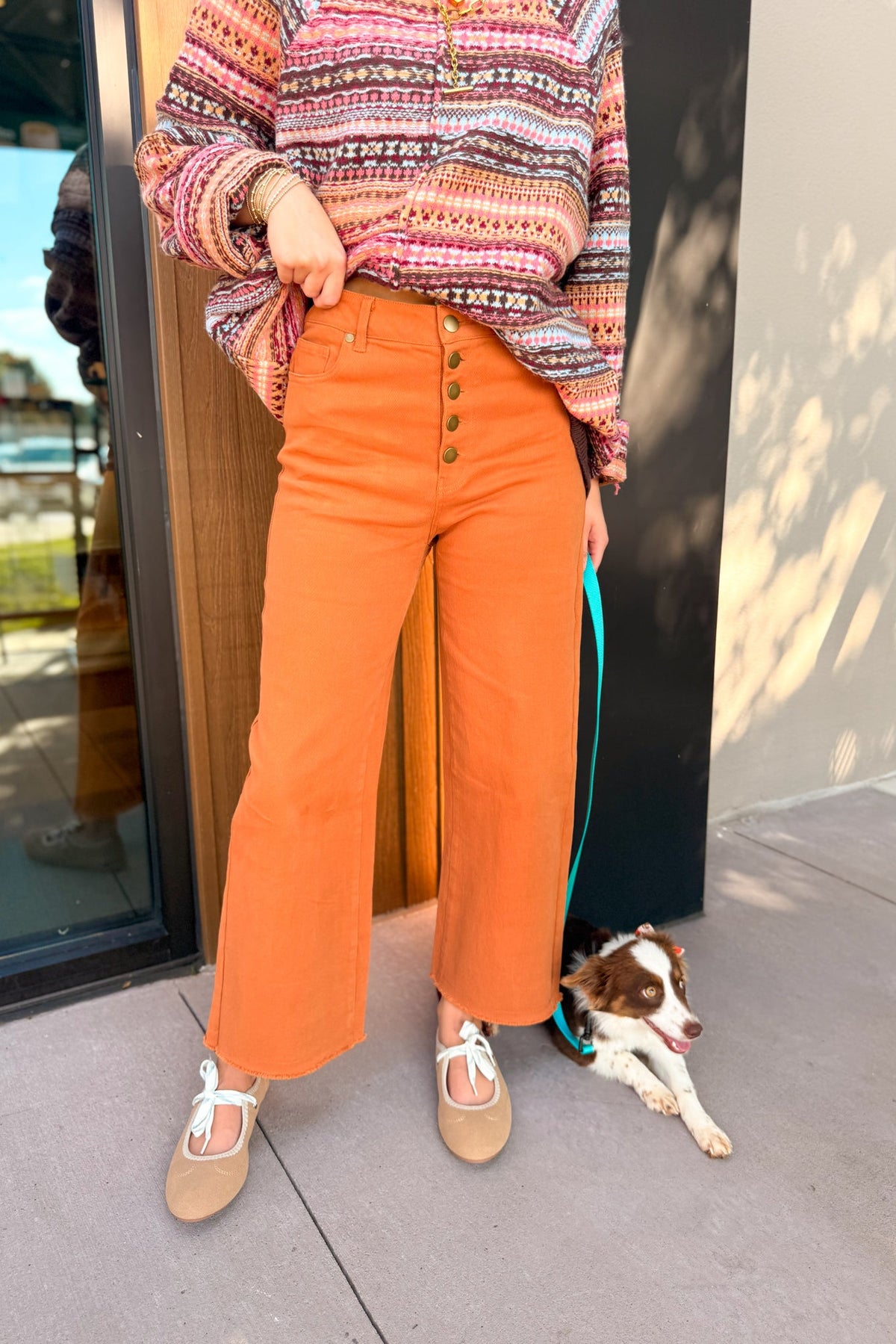 A person wearing bright orange wide-leg cropped pants with a button fly, styled with a multicolor patterned knit sweater and beige flat shoes, standing outdoors next to a brown and white dog on a teal leash.