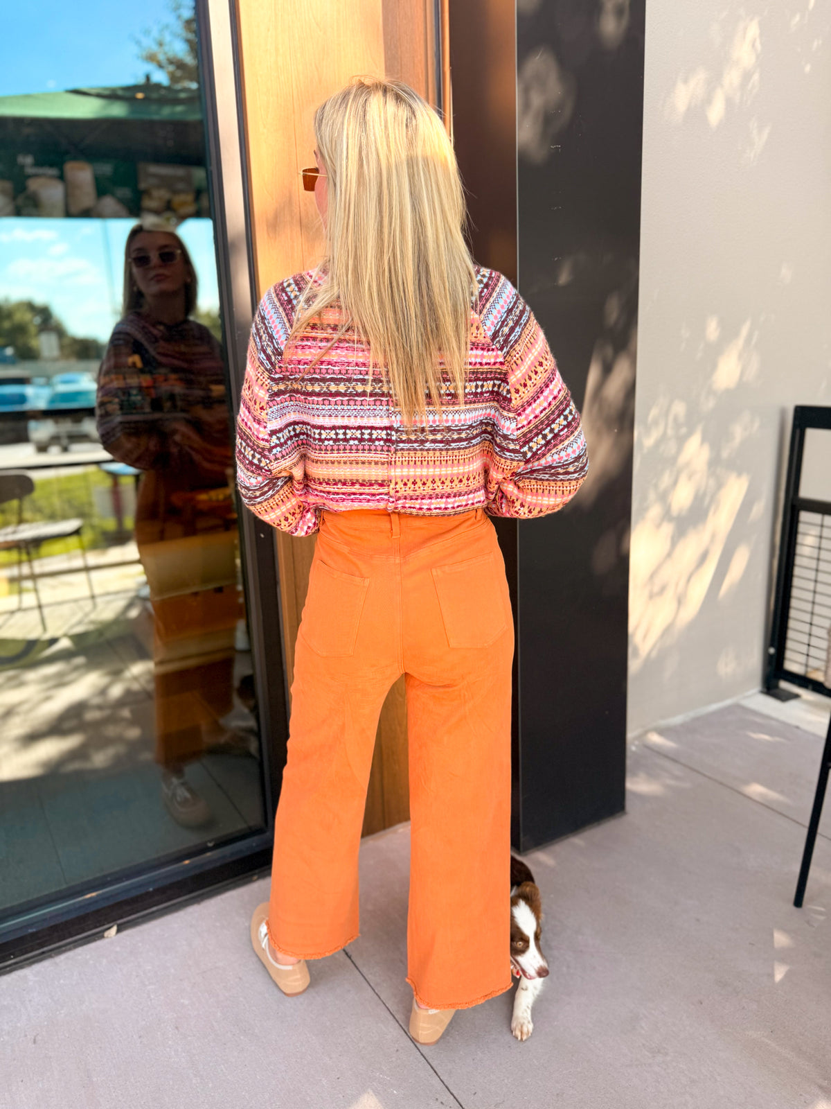 A woman stands with her back to the camera wearing a colorful patterned knit sweater and bright orange wide-leg pants, standing on a patio next to a glass door with a dog peeking out from underneath.